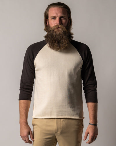 A man with a full beard and long hair wears the Dandy Del Mar Grenada Knit Baseball Tee - Tierra with tan pants, standing against a plain background.