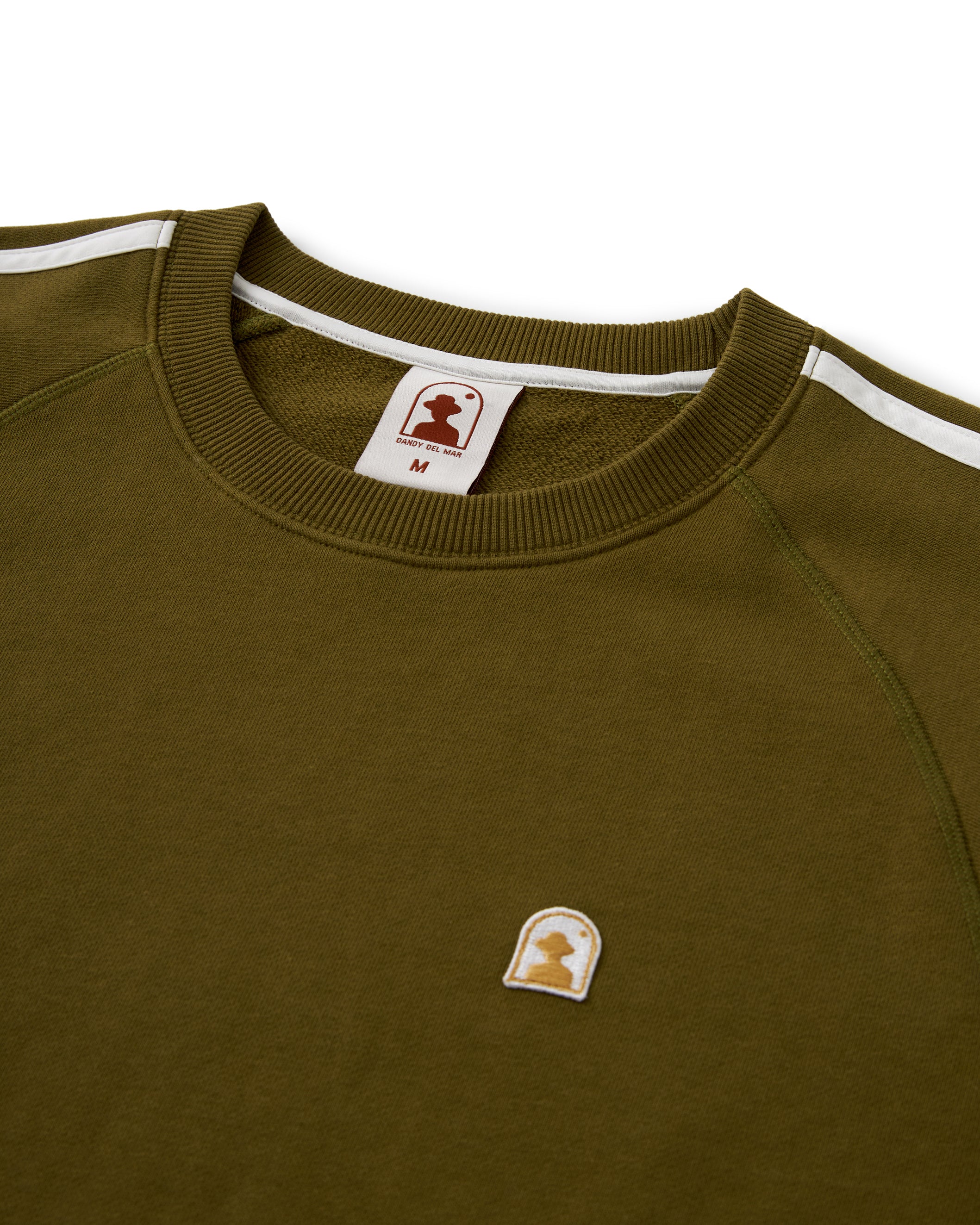 The Marseille French Terry Pullover - Arbequina by Dandy Del Mar is an olive green sweat top with raglan crew sleeves and white stripes on the shoulders. It features a small logo patch on the chest and has a size label marked "M." Crafted from French terry fabric, it offers ultimate comfort.