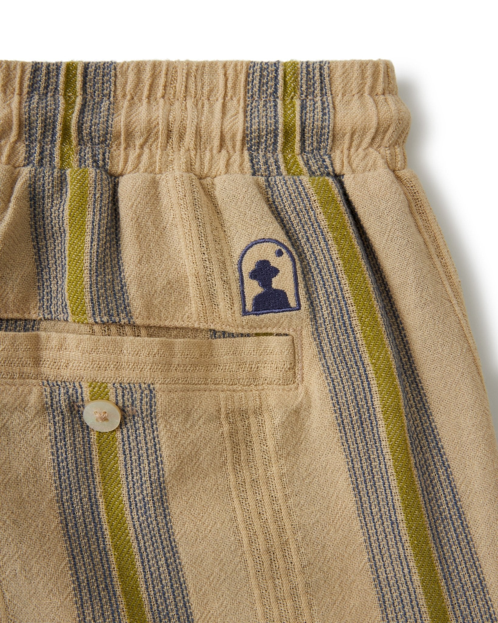 Close-up of The Corralejo Shorts - Ginger by Dandy Del Mar: beige striped crinkle gauze, elastic waistband, back pocket with button, and a small blue embroidered logo above the pocket. Summer essentials redefined.
