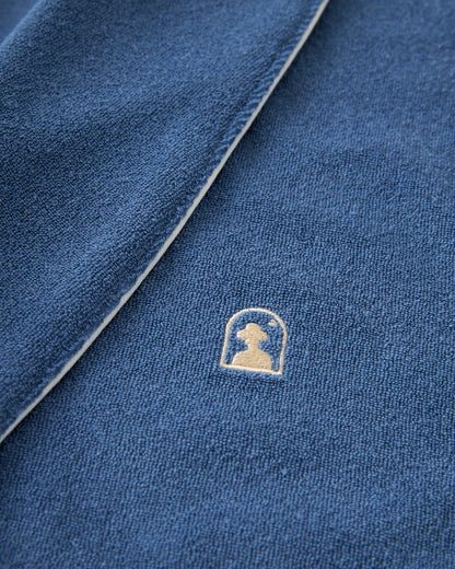 Close-up of The Tropez Robe - Annapolis by Dandy Del Mar, showcasing its ultra-soft blue fabric and a small embroidered logo of an arch and silhouette, reminiscent of premium terry cloth.