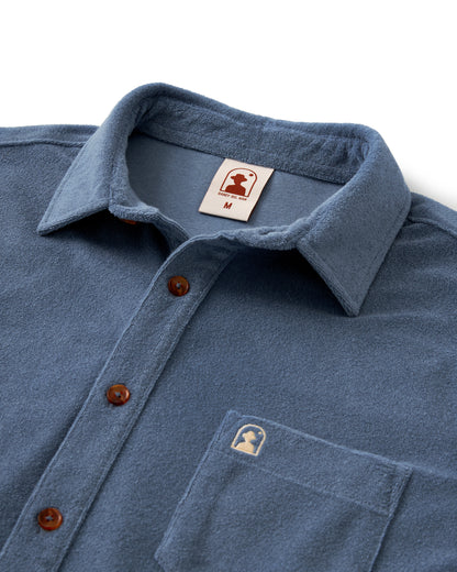 The Dandy Del Mar Tropez Shirt - Annapolis is a blue collared button-up with brown buttons, made from premium terry cloth. It includes a left chest pocket and a small logo tag, capturing the charm of the Côte d'Azur.