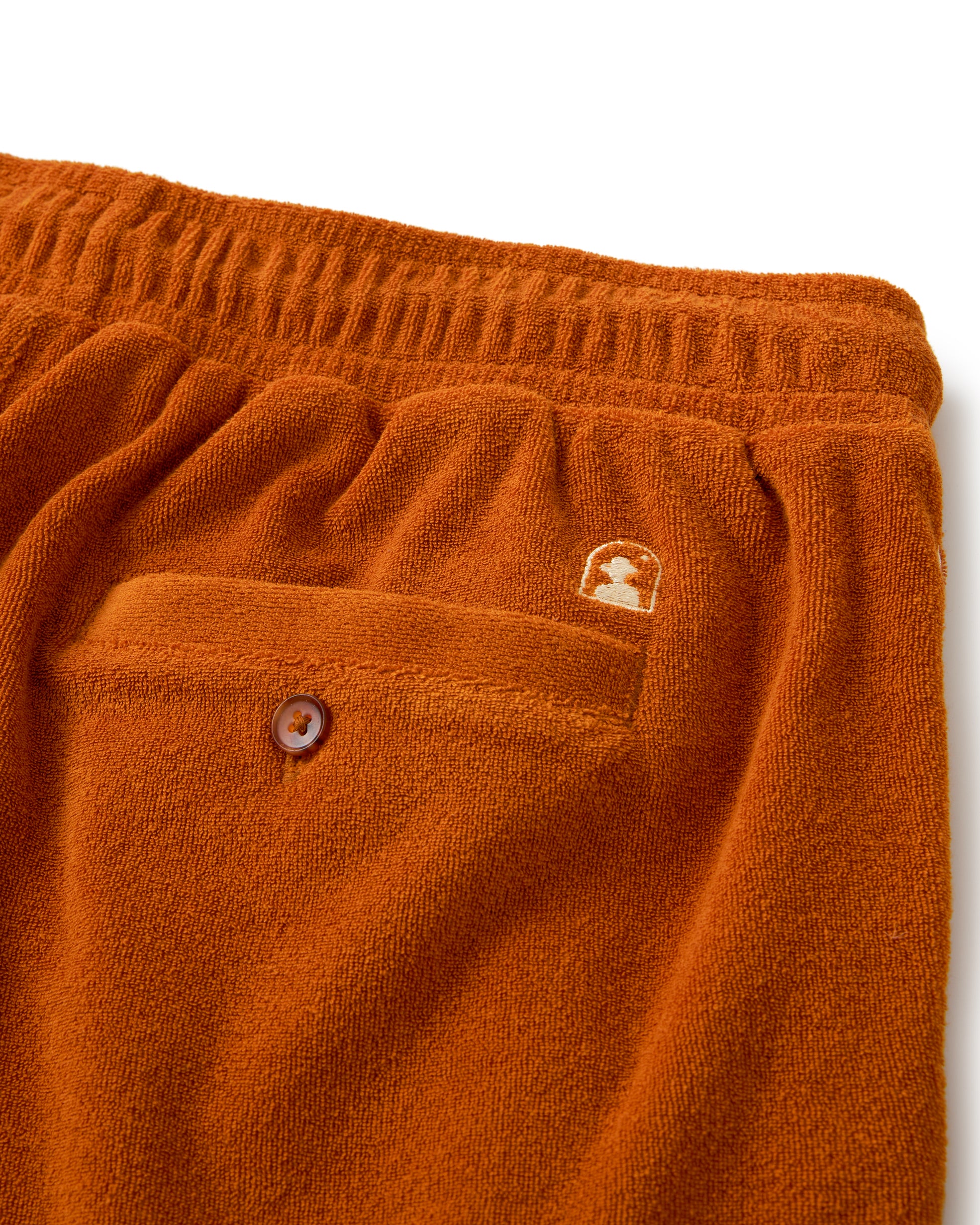 Close-up of The Tropez Short in Burnt Sienna by Dandy Del Mar, showcasing orange textured fabric, an elastic waistband, buttoned back pocket, and a small embroidered emblem above the pocket. Ideal for sun-soaked days on the Côte d'Azur.