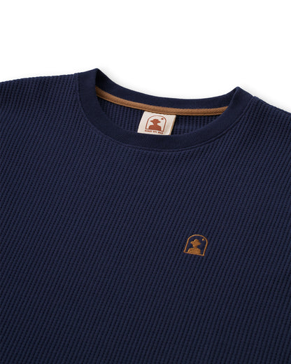 Close-up of The Cannes Long Sleeve Shirt - Luxe Navy featuring a waffle-knit texture, round neck, and an embroidered Dandy Del Mar logo with a silhouette of a person under an arch on the left chest. This long-sleeve tee from Dandy Del Mar perfectly blends style and comfort.