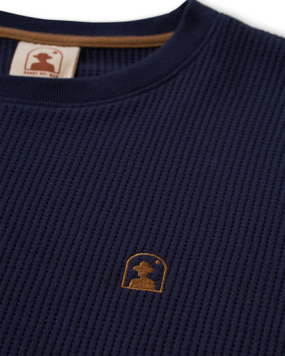 Close-up of The Cannes Long Sleeve Shirt - Luxe Navy by Dandy Del Mar, showcasing its waffle knit fabric and a small embroidered logo on the chest, featuring a head inside an arched outline. A neck tag is also visible at the collar.