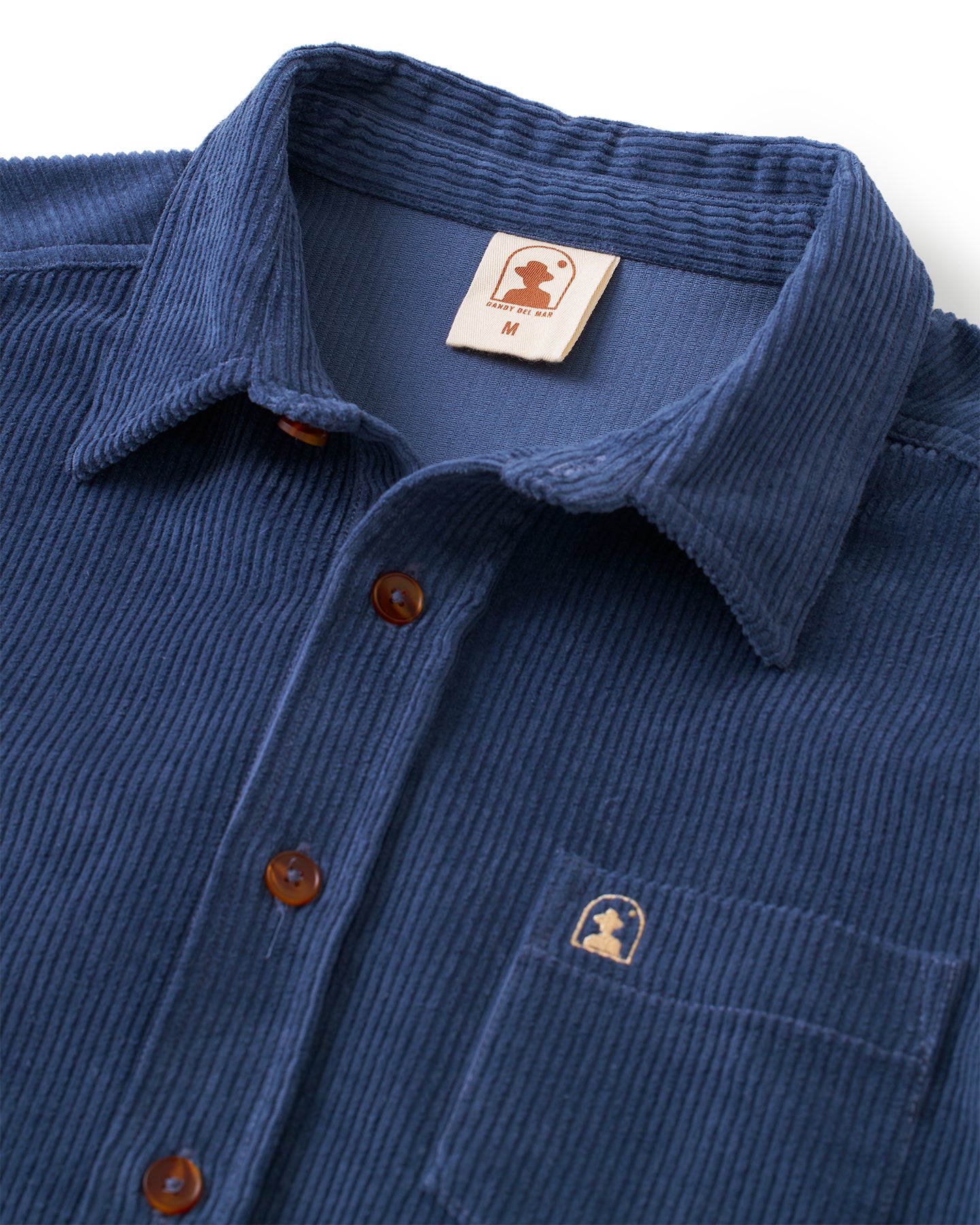 Close-up of the blue Corsica Corduroy Shirt - Moontide by Dandy Del Mar in an 8-wale corduroy with a tailored relaxed fit, featuring a collar, brown buttons, and a logo tag above the left chest pocket.