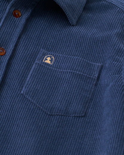 Close-up of The Corsica Corduroy Shirt - Moontide by Dandy Del Mar, showcasing an 8-wale corduroy texture and a front pocket with a small yellow logo embroidered near the top. The shirt features brown buttons and boasts a tailored relaxed fit.