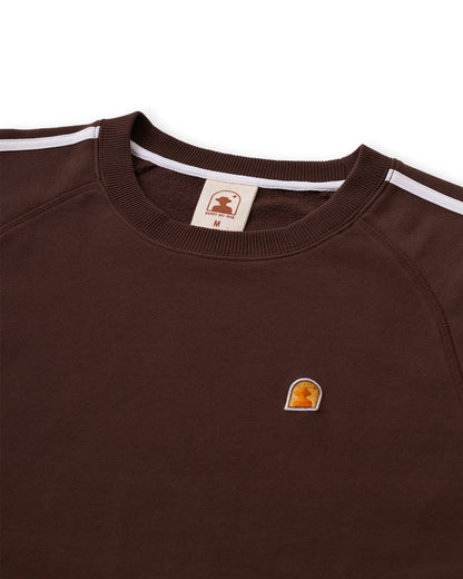 The Marseille French Terry Pullover - Carajillo by Dandy Del Mar is a brown crewneck sweater with white shoulder stripes, featuring a small embroidered logo on the chest, a branded tag on the collar, and a ribbed neckband crafted from soft French terry fabric.