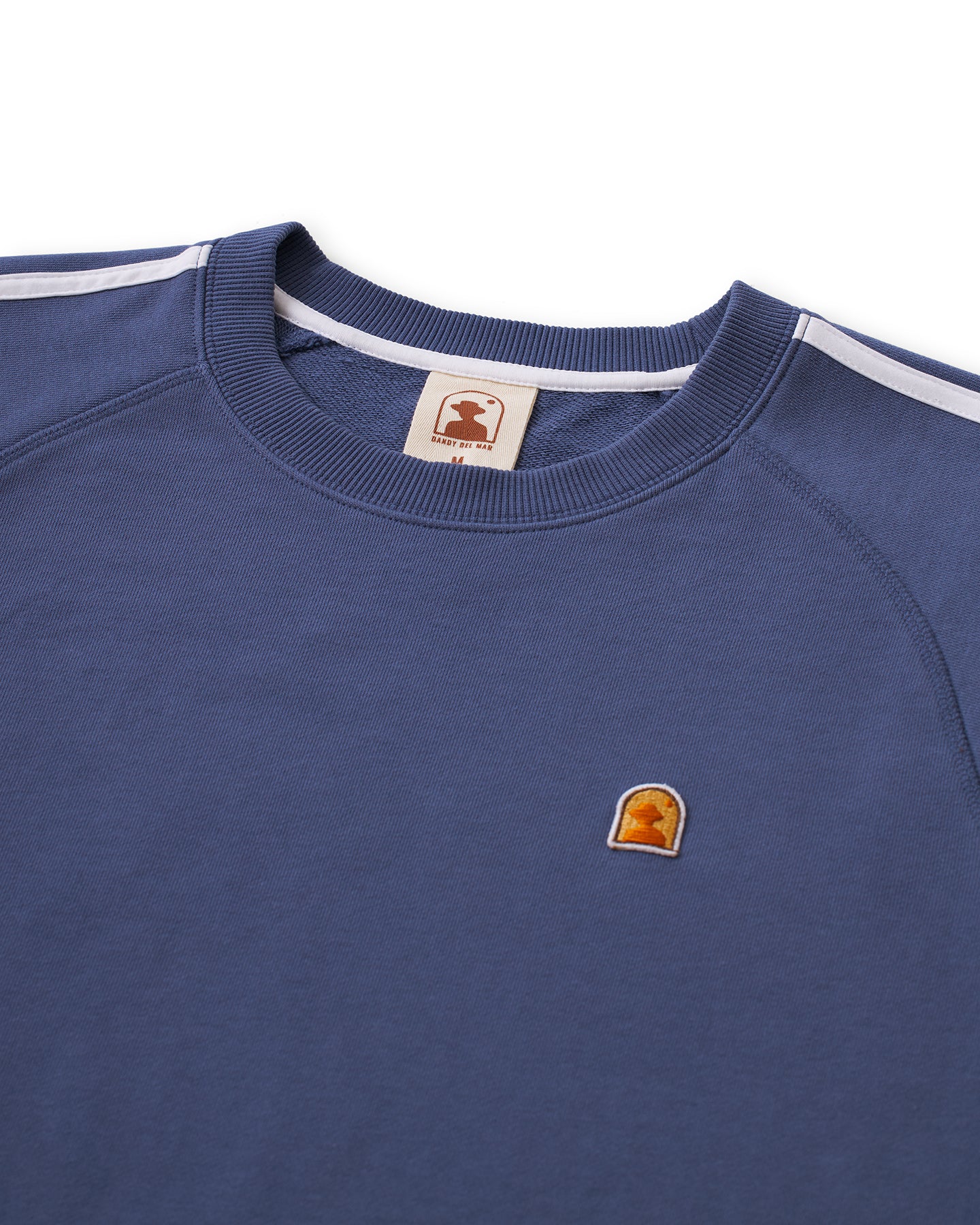 A close-up of the Marseille French Terry Pullover - Moontide by Dandy Del Mar, showcasing a blue French Terry sweatshirt with white stripes on the sleeves and a small embroidered logo patch on the chest depicting a silhouette of a head.