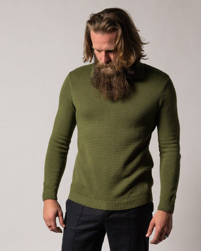 A man with long hair and a full beard looks down, wearing Dandy Del Mar’s The Sebastian LS Polo Shirt in Arbequina and dark pants against a plain background.