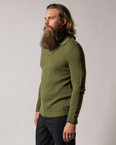A man with long hair and a full beard wears Dandy Del Mar’s The Sebastian LS Polo Shirt in Arbequina and dark pants, standing against a plain light background.