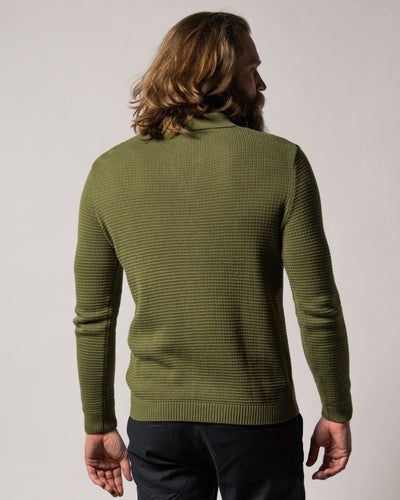 A man with long hair stands facing away, wearing the Dandy Del Mar Sebastian LS Polo Shirt in Arbequina and dark pants against a neutral background, highlighting the knitwear collection.