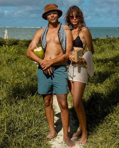 Two people stand barefoot on a grassy beach path, each holding a drink and wearing sunglasses. One wears Dandy Del Mar’s The Ventura Volley Shorts - Acqua, while the ocean glimmers in the background amidst their stylish look.