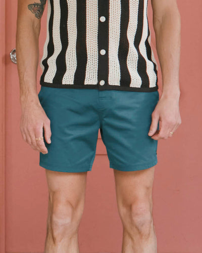 A person is wearing a black and white striped crochet sleeveless top and Dandy Del Mar's The Mallorca Swim-Walk Shorts in Tempest Blue, featuring adjustable brass fasteners, standing before a pink wall with only the torso and legs visible.