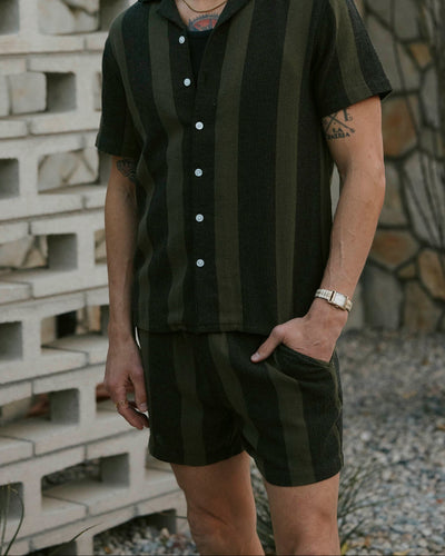 A man stands outside wearing Dandy Del Mar’s The Palma Shorts in Onyx, a relaxed fit short-sleeve, button-up and matching shorts set featuring dark green and black yarn dyed stripes, with one hand in his pocket.