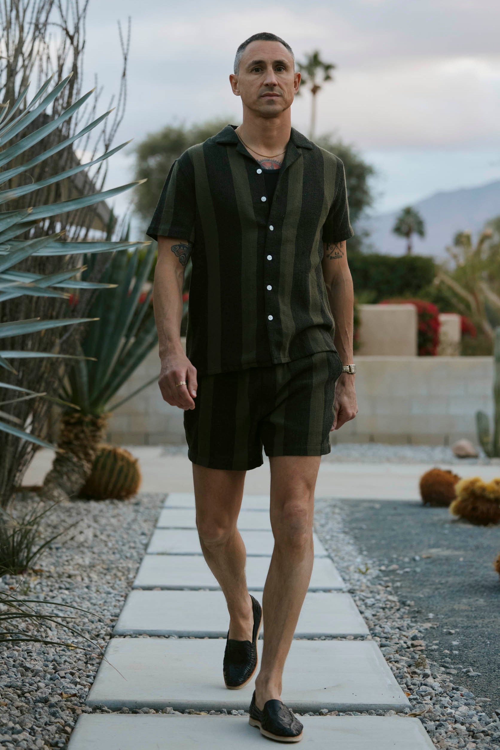 A person strolls along a stone path in a desert garden, sporting The Palma Shirt in Onyx by Dandy Del Mar, accompanied by shorts. Behind them are desert flora and a cloudy sky.
