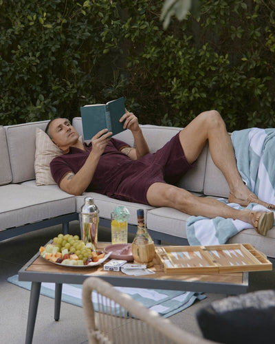 A man in Dandy Del Mar’s The Rio Velour Shorts - Bordeaux relaxes on an outdoor sectional, reading a book with snacks, drinks, and a backgammon board on a nearby table.