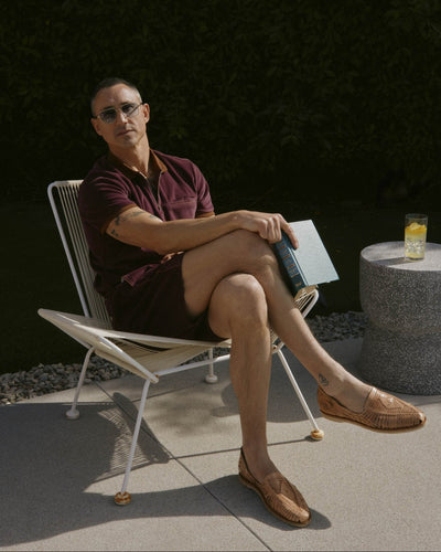 A person in Dandy Del Mar’s The Rio Velour Shorts in Bordeaux and sunglasses sits on a patio chair with legs crossed, holding a book. A drink rests on a nearby round stone table.