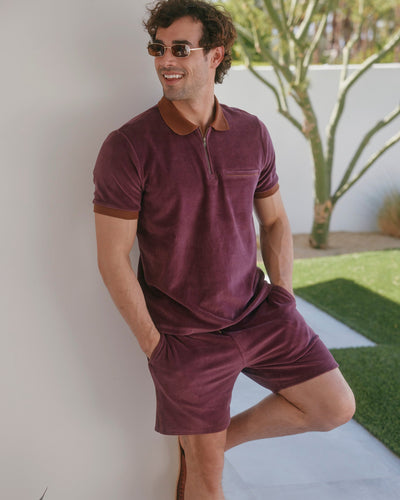 A man in Dandy Del Mar's The Rio Velour Shorts - Bordeaux stands casually against a white wall outdoors, wearing matching maroon sunglasses and shirt, with greenery in the background.