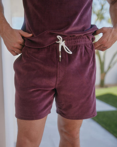 A man stands outdoors wearing The Rio Velour Shorts in Bordeaux by Dandy Del Mar, with hands on the elastic waistband—ideal for men's leisurewear.