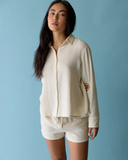 A woman stands against a blue background wearing Dandy Del Mar’s cream-colored Sevilla Top - Scallop with matching shorts.
