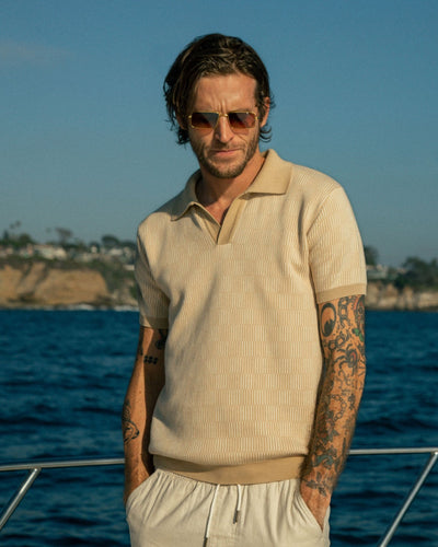 Tattooed man in sunglasses, The Sebastian Knit Polo Shirt - Shell by Dandy Del Mar, and light shorts stands on a boat with the ocean and rocky coastline in the background.