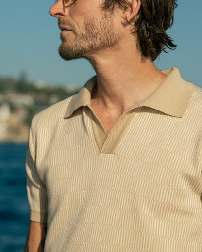 Wearing Dandy Del Mar's The Sebastian Knit Polo in Shell, a person gazes into the distance by the seaside on a sunny day, epitomizing Italian knit elegance.