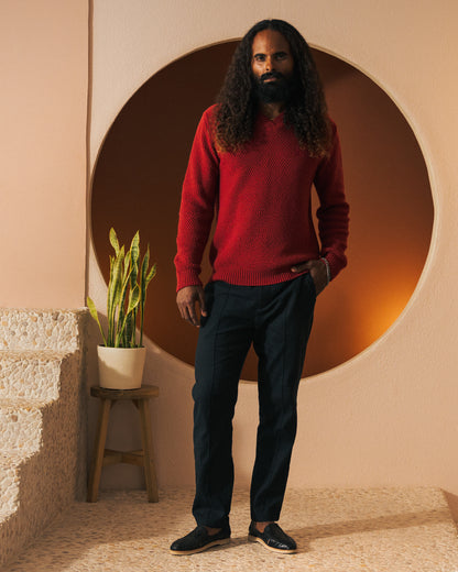 A person with long curly hair stands indoors, wearing "The Algiers Knit Sweater - Fuego" by Dandy Del Mar and dark pants. A small plant in a pot is on a stool to their left. Circular art decor adorns the wall behind them.