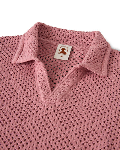 A close-up of The Antibes Crochet Polo Shirt - Dawn by Dandy Del Mar, featuring a pink knit, V-neck, diamond pattern, collared design, and a visible size M tag.
