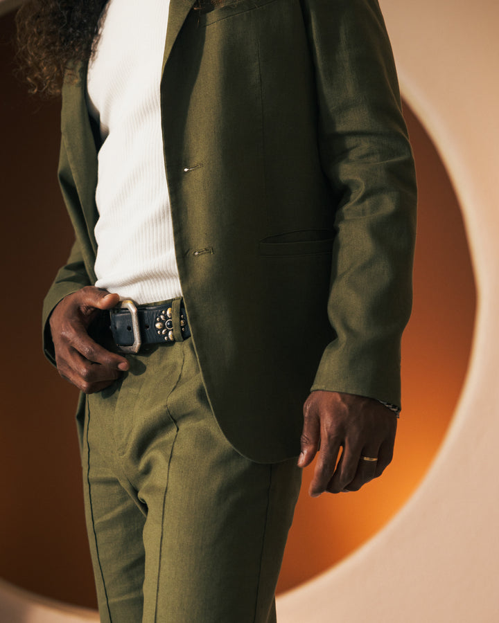 A person wearing The Brisa Linen Blazer - Berdea by Dandy Del Mar in a green hue, paired with a white shirt, is shown from the chest to thigh level, with one hand placed near the belt buckle.