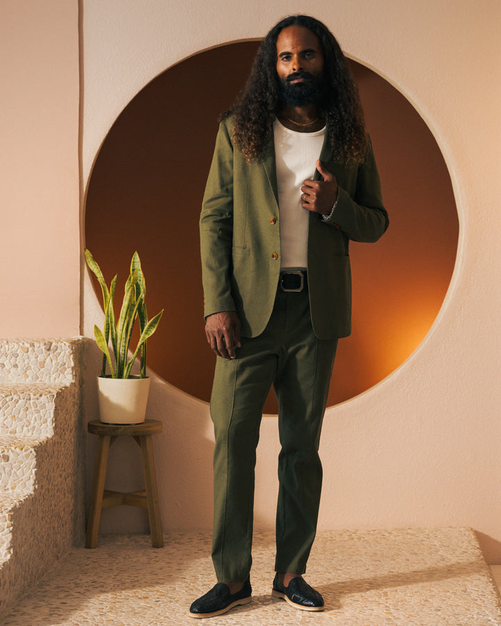 A person with long curly hair and a beard is standing in front of a circular backdrop, wearing The Brisa Linen Blazer - Berdea by Dandy Del Mar over a white shirt, next to a potted plant on a stool.