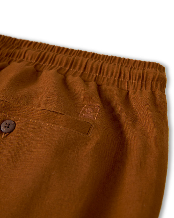 Close-up of a pair of The Brisa Linen Pant - Sedona by Dandy Del Mar in brown, featuring an elastic waistband and a buttoned pocket. A small embroidered logo is visible above the pocket, making these vacation pants a stylish and practical choice.