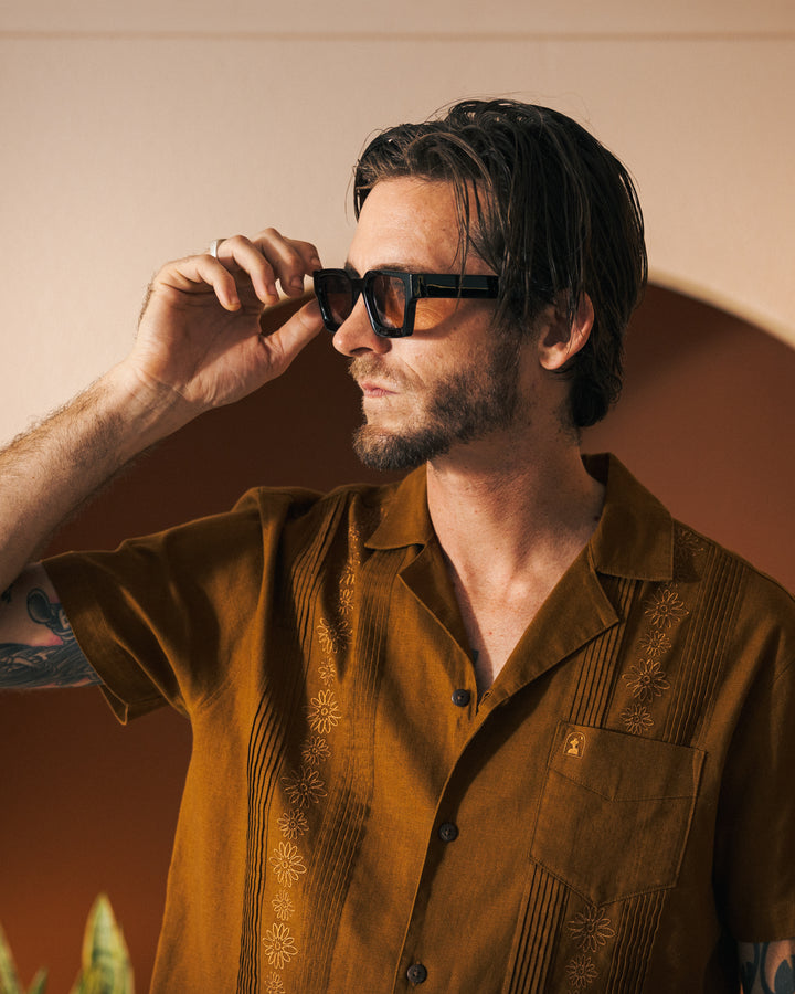 A man with shaggy hair and a beard, wearing dark sunglasses and a brown Sedona Brisa Linen Shirt from Dandy Del Mar, adjusts his glasses with one hand. He stands in a warmly lit room with a plant in the background.