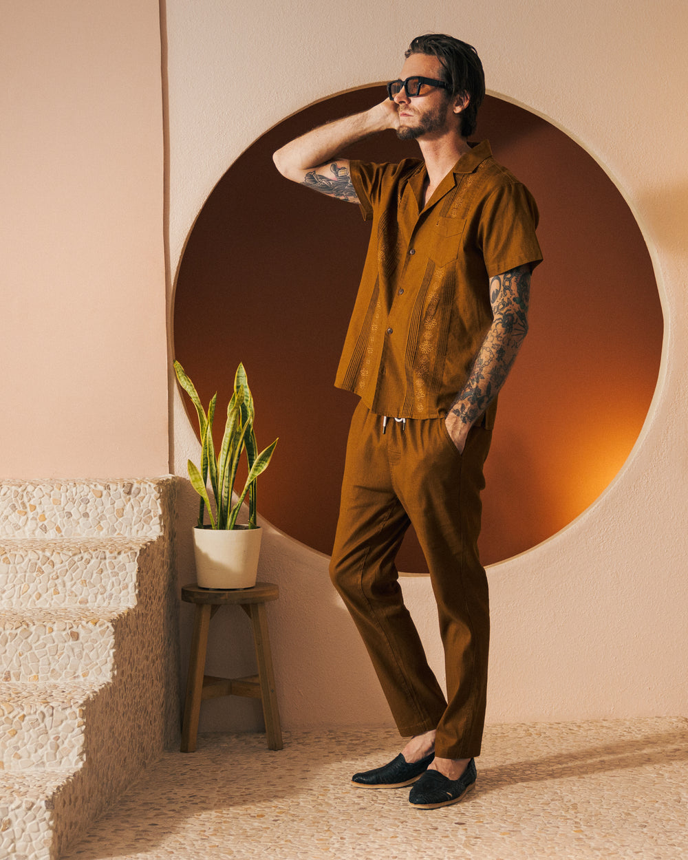 A man in a brown short-sleeve shirt and matching "The Brisa Linen Pant - Sedona" from Dandy Del Mar stands beside a stairway, posing with one hand on his temple. A plant in a pot is on the short stool beside him.