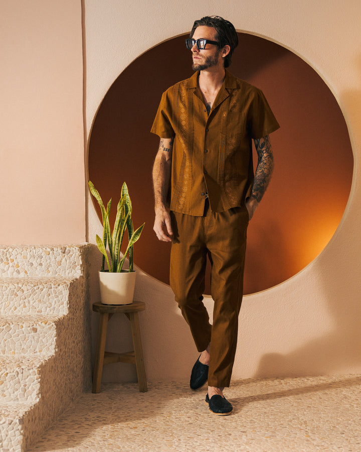 A man in sunglasses and Dandy Del Mar's The Brisa Linen Pant in Sedona stands near a staircase with a potted plant on a stool, in front of a circular orange backdrop.