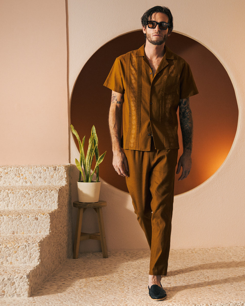 A person wearing dark sunglasses and The Brisa Linen Shirt in Sedona by Dandy Del Mar, adorned with gardenia floral embroidery, along with matching pants featuring coconut shell buttons, stands indoors next to a plant on a stool and a staircase with pebble-textured steps.