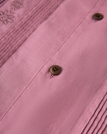 A close-up of The Brisa Linen Shirt - Dawn by Dandy Del Mar shows pink fabric detailing with two brown buttons, vertical pleats on the left side, and floral embroidery in the upper left corner.