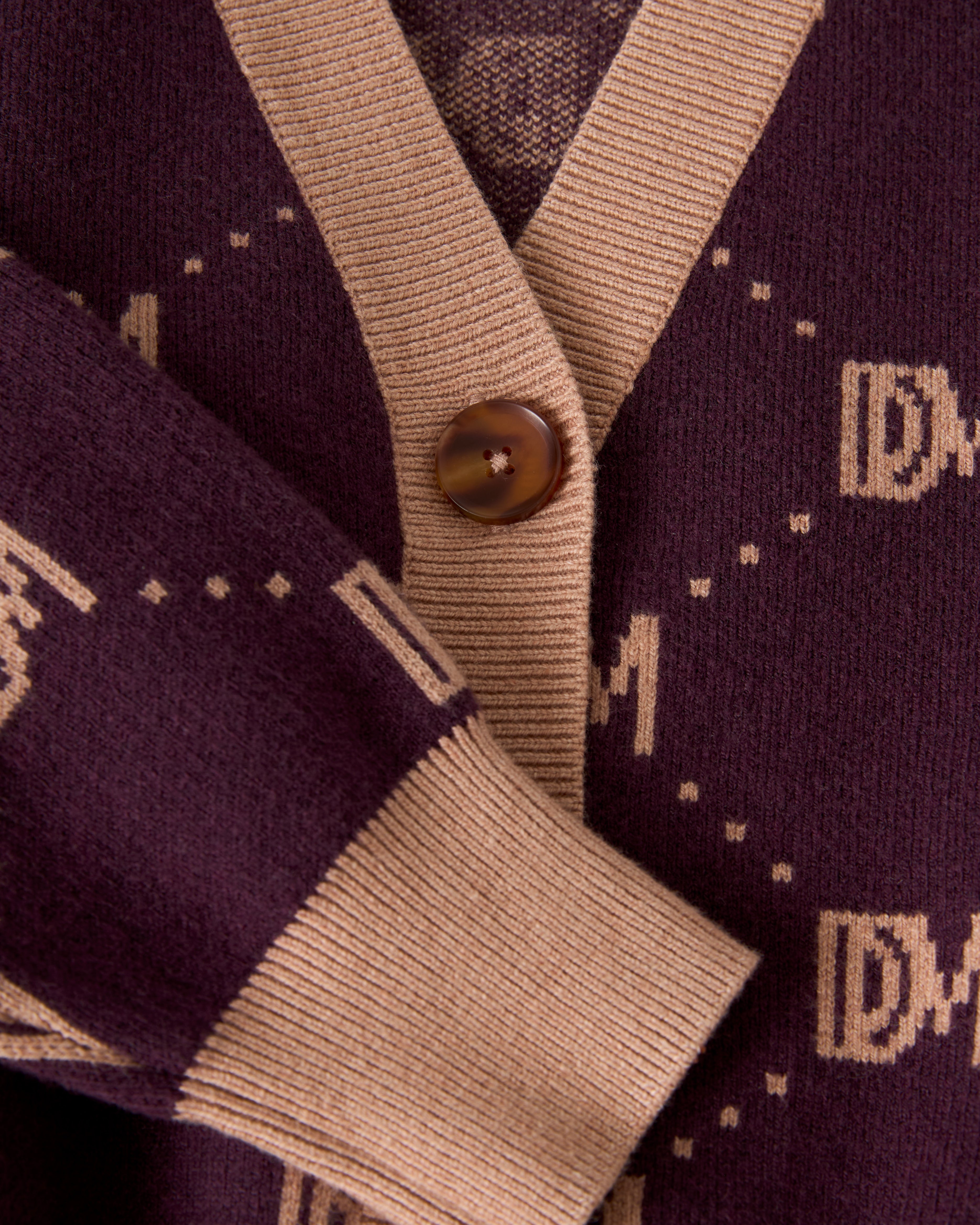 Close-up of The Canela Cardigan - Amethyst from Dandy Del Mar, showcasing a maroon cotton design with beige trim and button, adorned with a stylish monogram pattern.