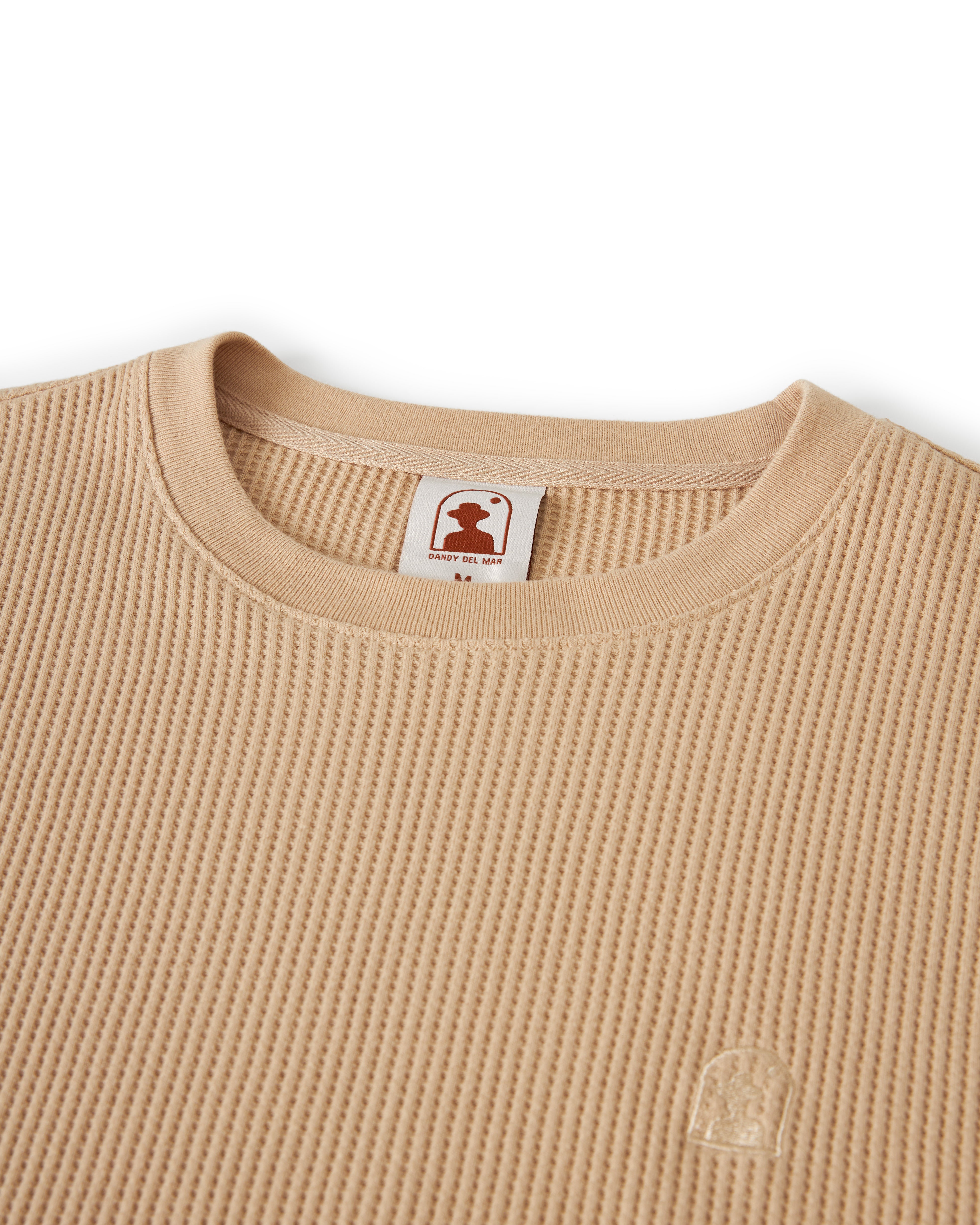 Close-up of The Cannes Long Sleeve Shirt in Truffle by Dandy Del Mar, a beige waffle-knit shirt made from 100% cotton, featuring a small logo emblem on the bottom right and a neck tag reading "Heavy Big Man.