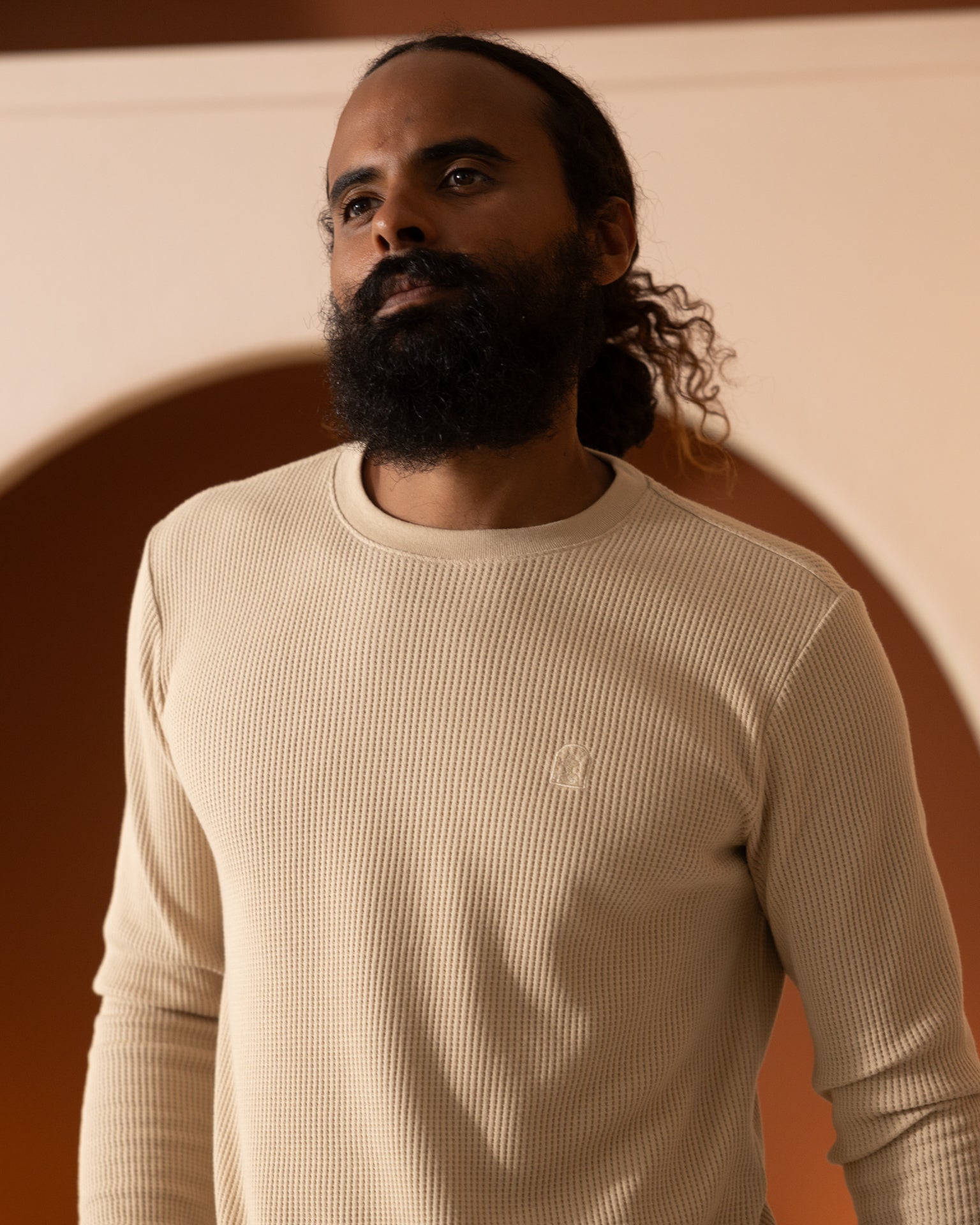 A man with a beard and curly hair tied back, wearing The Cannes Waffle Knit Shorts in Truffle by Dandy Del Mar and a beige long-sleeve ribbed shirt made from 100% cotton, stands in an indoor setting with arched walls.