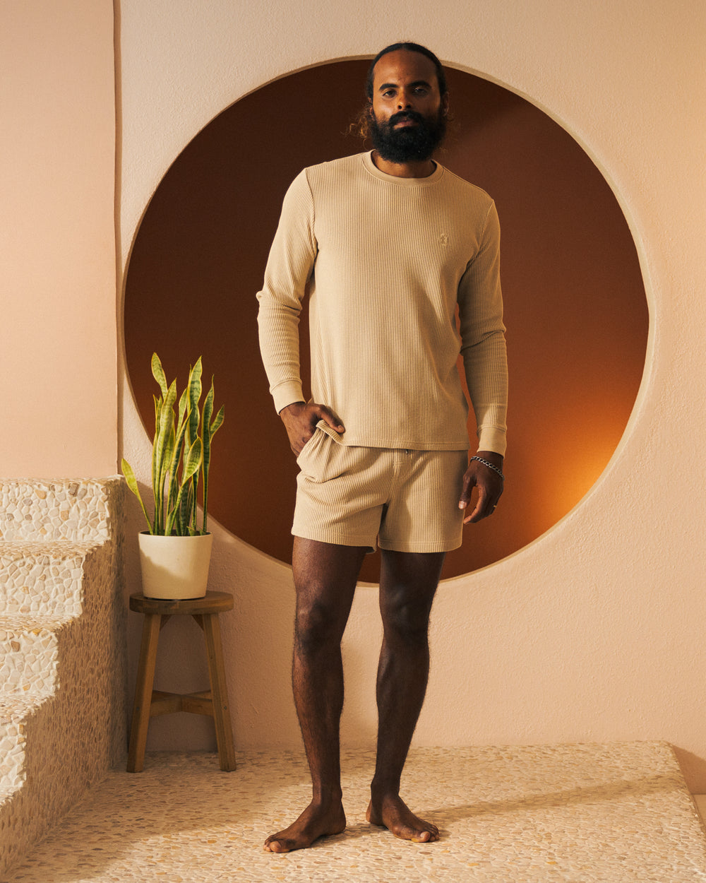 A man with a beard and long hair is standing indoors wearing a beige long-sleeve shirt made from waffle knit fabric and Dandy Del Mar's The Cannes Waffle Knit Shorts - Truffle. He is barefoot and standing next to a potted plant on a stool against a circular orange background.