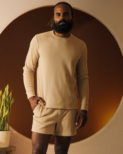 A bearded man stands before a round brown backdrop, wearing Dandy Del Mar’s The Cannes LS Waffle Knit Shirt in Truffle and matching shorts with an embroidered logo. A potted plant is visible to his left.