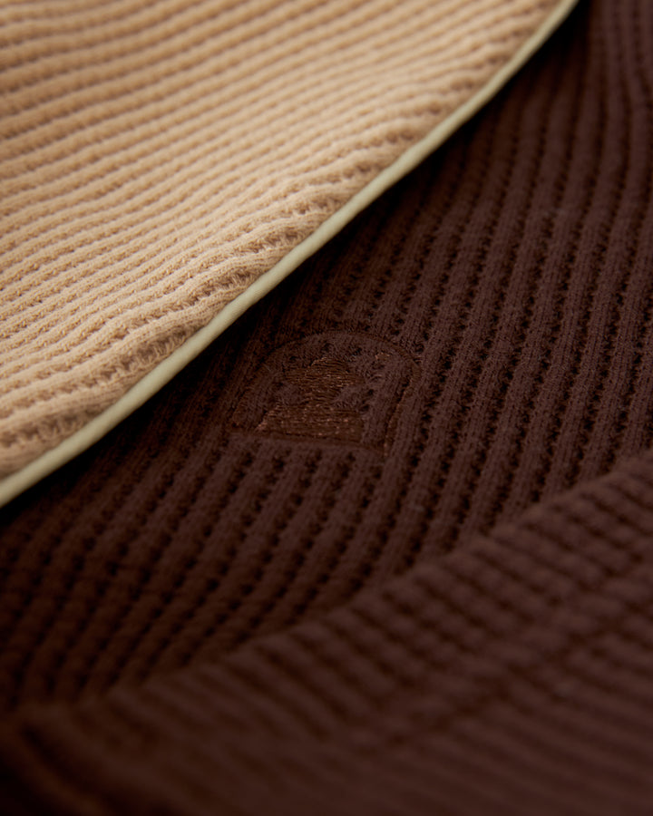 Close-up of beige and brown waffle knit fabrics overlapping each other with a small logo embossed in the brown fabric, resembling the elegant texture of The Cannes Waffle Knit Robe - Carajillo by Dandy Del Mar.