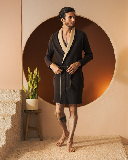 A man stands on a terrazzo floor, dressed in The Cannes Waffle Knit Robe - Carajillo by Dandy Del Mar, showcasing luxurious comfort with its cream-collared brown waffle knit design, in front of a round backdrop. A potted plant is placed on a stool beside him.