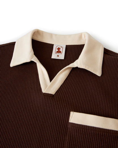 Close-up of the Dandy Del Mar Cannes Waffle Knit Shirt - Carajillo, showcasing its dark brown hue with a white collar and trim, a V-neck, and a pocket adorned with a white band. The waffle knit fabric includes a label featuring a figure and the letter 'M'.