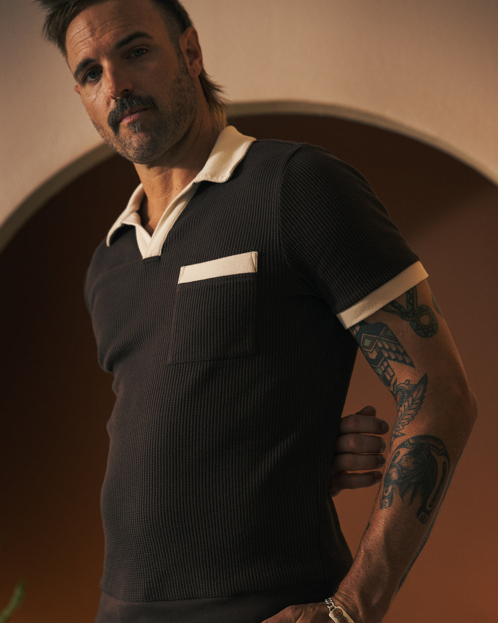 A man with tattoos on his left arm and wearing a short-sleeve dark Cannes Waffle Knit Shirt - Carajillo by Dandy Del Mar with a white collar and pocket stands indoors in front of an arched doorway, looking directly at the camera.