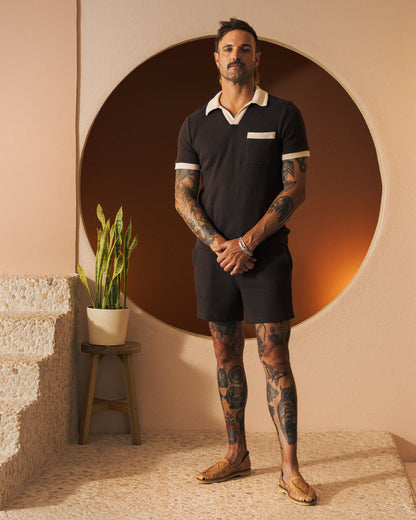 A man with arm and leg tattoos stands in a room, wearing a dark short-sleeved Cannes Waffle Knit Shirt - Carajillo from Dandy Del Mar, paired with shorts. He is posed by a round wall feature and a plant on a stool.