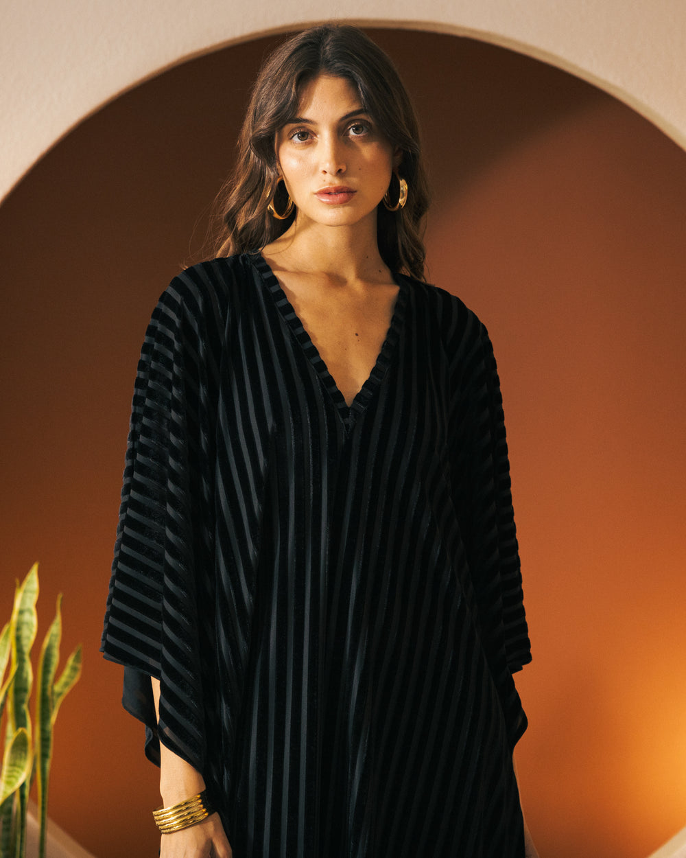 A person with long wavy hair wearing The Marsala Kaftan - Onyx by Dandy Del Mar and large hoop earrings stands indoors in front of an archway and brown background.