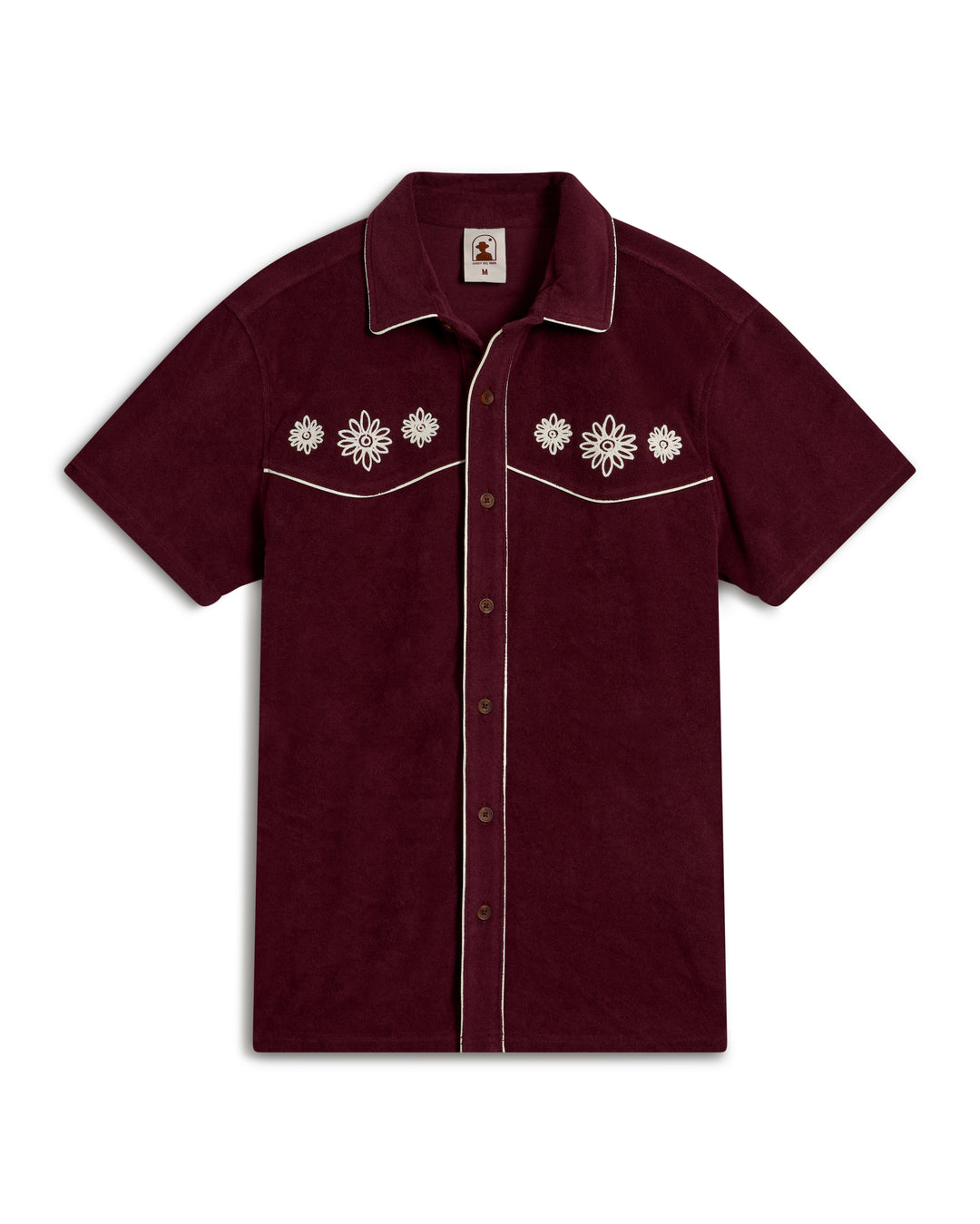 The Dandy Del Mar Gaucho Terry Cloth Shirt in Bordeaux is a maroon short-sleeved shirt with white floral embroidery on the chest, a button-down front, and white piping along the edges.