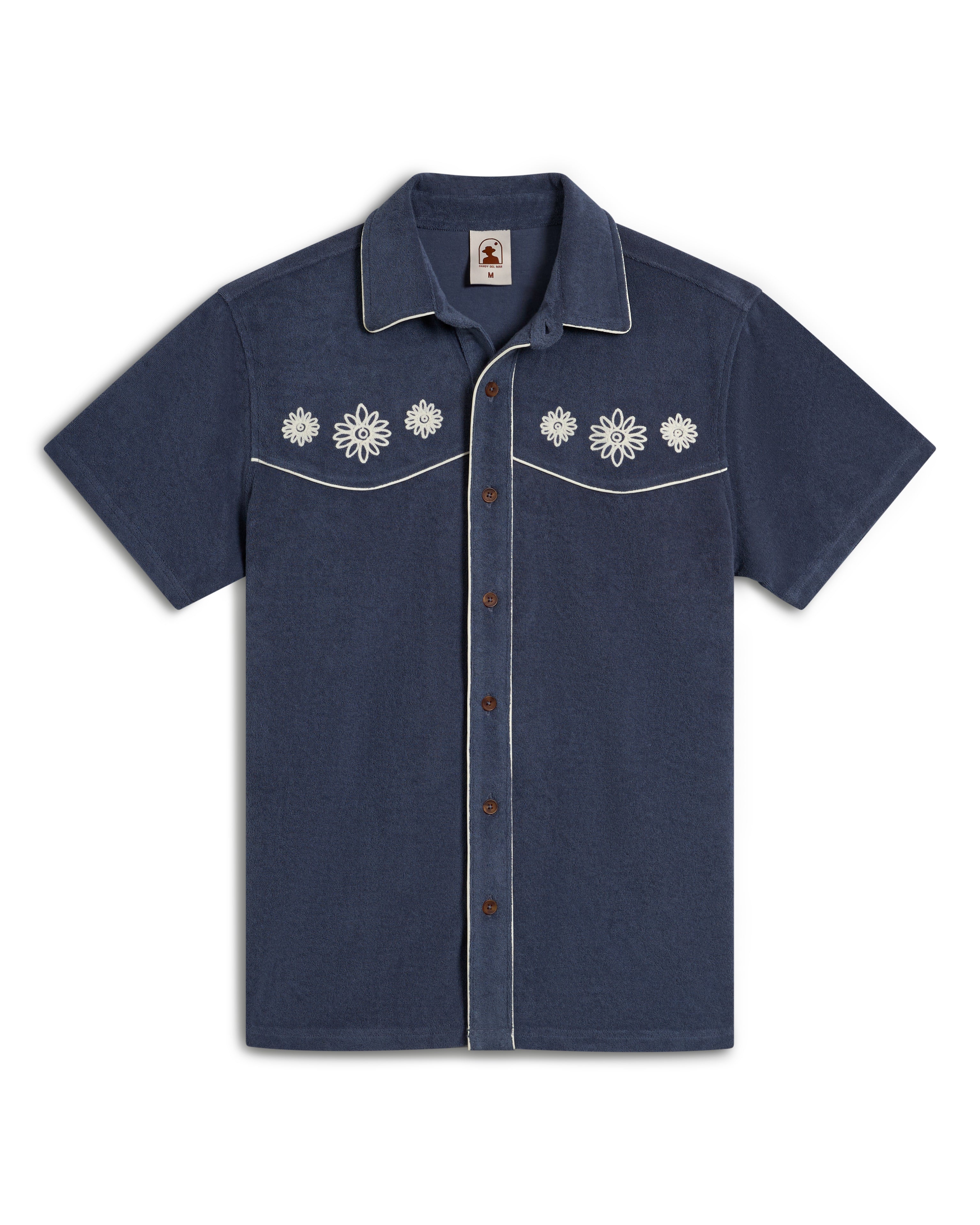 The Gaucho Shirt - Moontide by Dandy Del Mar is navy blue with white floral embroidery on chest pockets and white piping on edges, offering an Equatorial Cowboy style with brown front buttons.