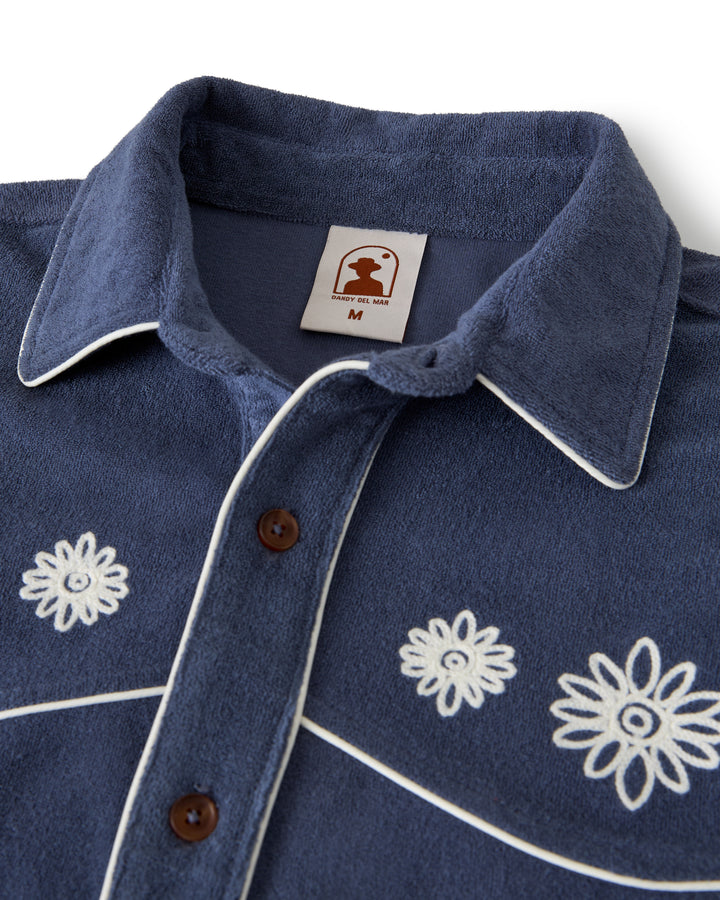 The Gaucho Shirt - Moontide by Dandy Del Mar features a blue texture with white floral embroidery and trim, brown buttons, and a logo tag with "M" size, perfect for that Equatorial Cowboy style.